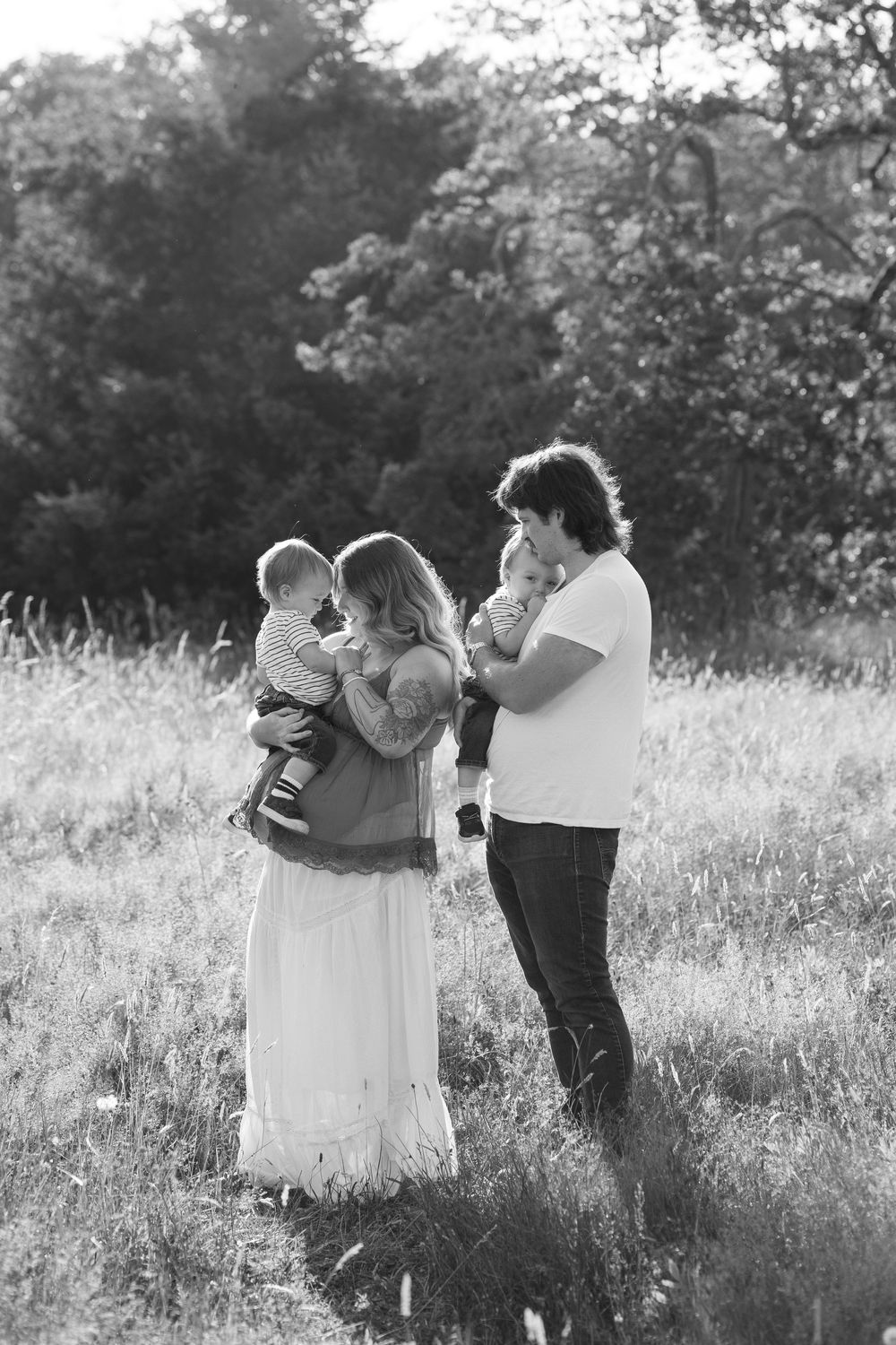 Black and white photo of a couple holding two babies in a grassy field with trees in the background.