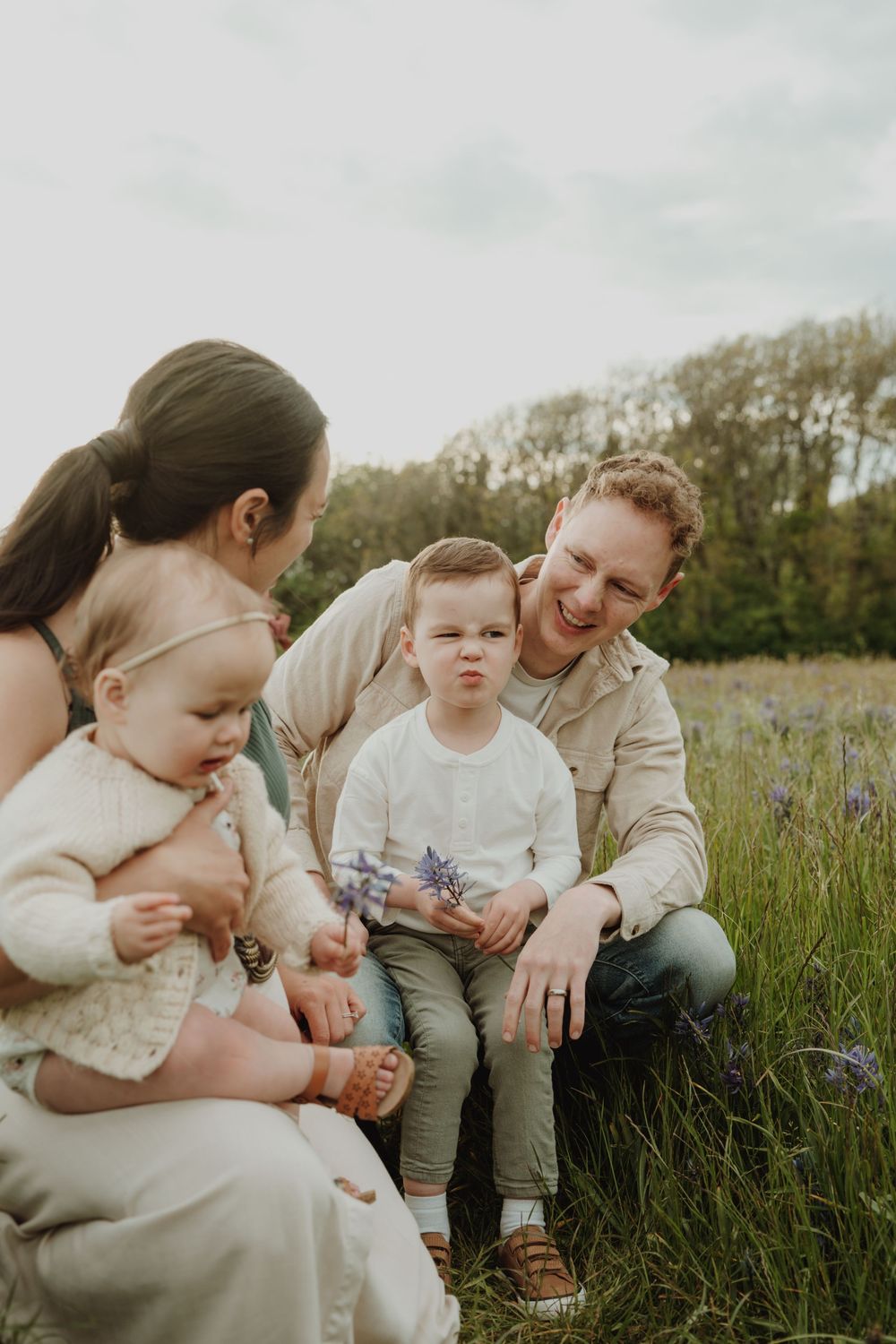 Family of four sits in a wildflower field, parents smiling at toddler boy holding purple flowers. Victoria, BC, Vancouver Island.