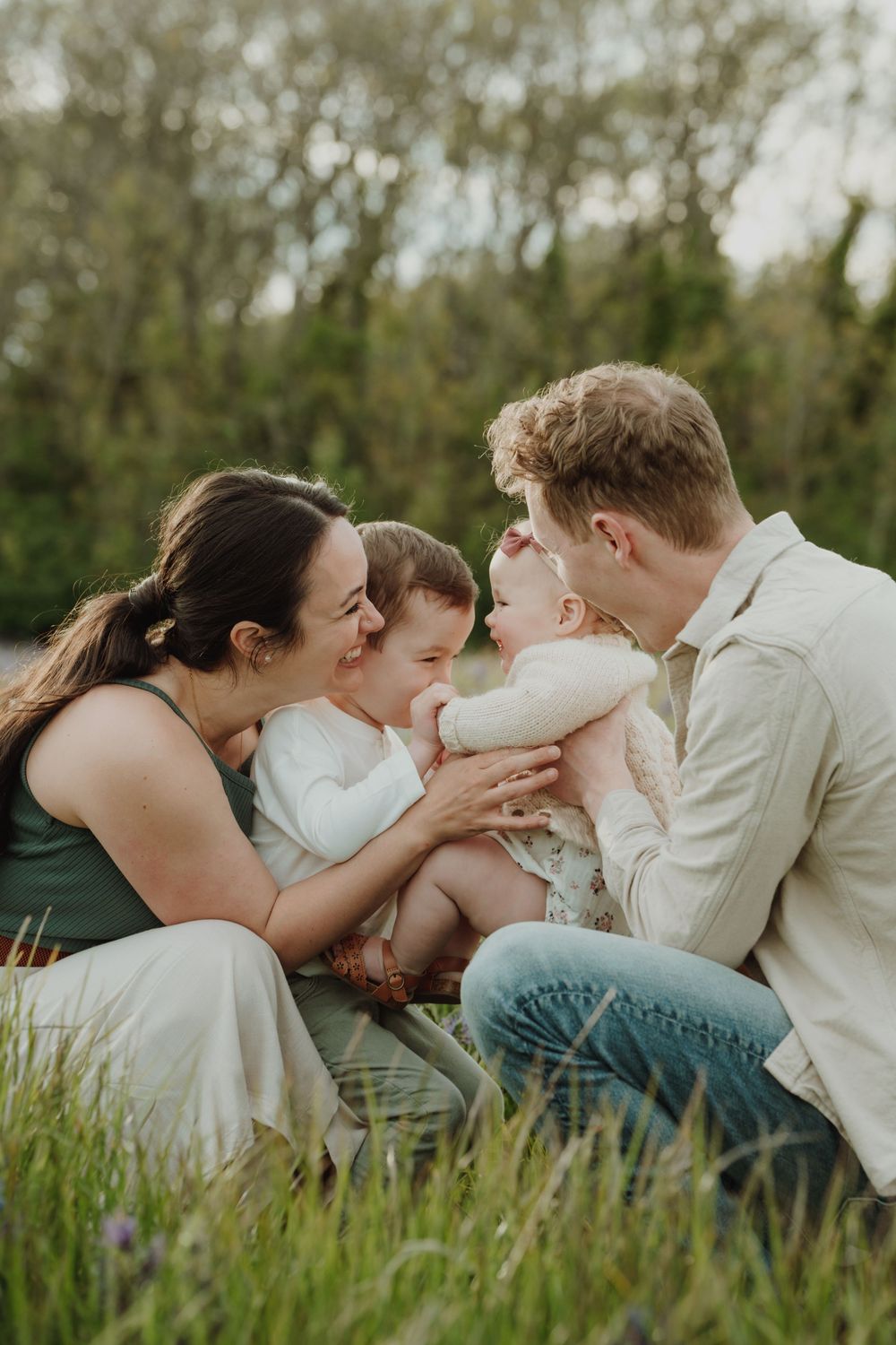 Loving parents lean in to kiss their two young children while sitting in green grass. Victoria, BC, Vancouver Island.