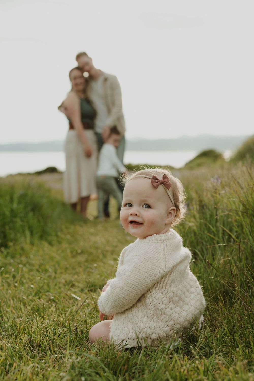 Smiling baby girl in cream knit sweater sits in grass with family embracing behind her. Victoria, BC, Vancouver Island.