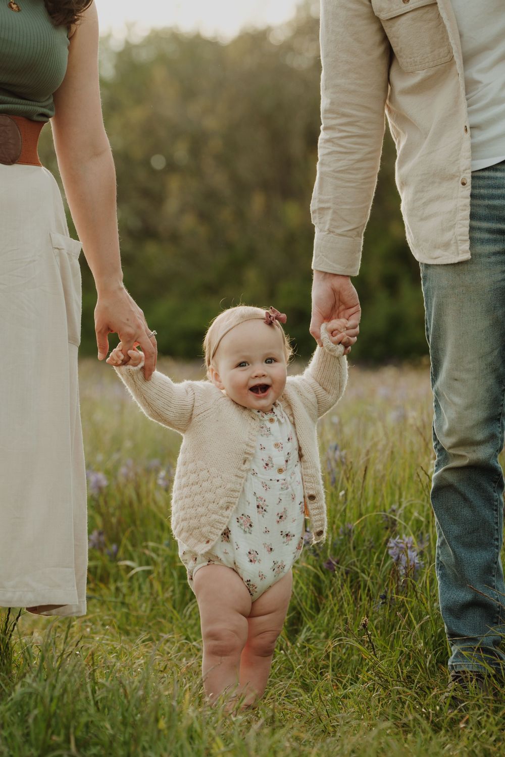 Baby girl in floral romper stands holding parents hands in a wildflower meadow at sunset. Victoria, BC, Vancouver Island.
