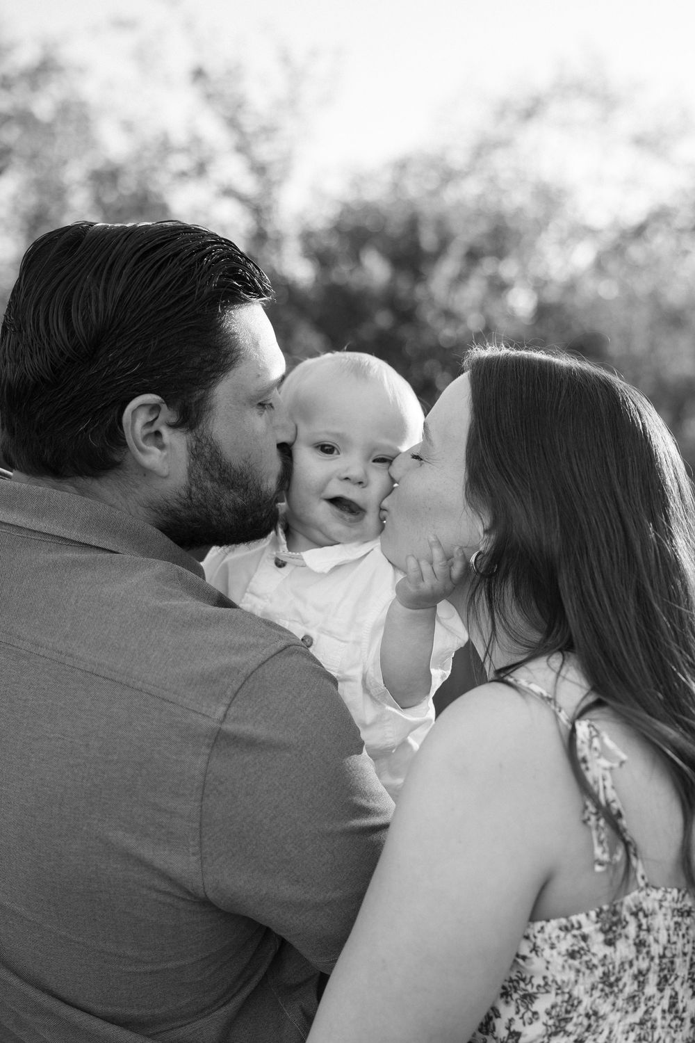 Black and white photo of parents kissing their smiling baby outdoors in a heartwarming family portrait.
