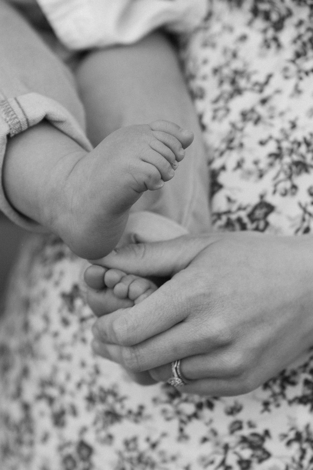 Black and white photo of an adult cradling tiny newborn baby feet, wearing a floral dress and diamond ring.