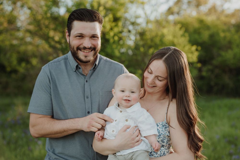 Smiling couple holding their happy baby outdoors in a lush green park during a family portrait session.