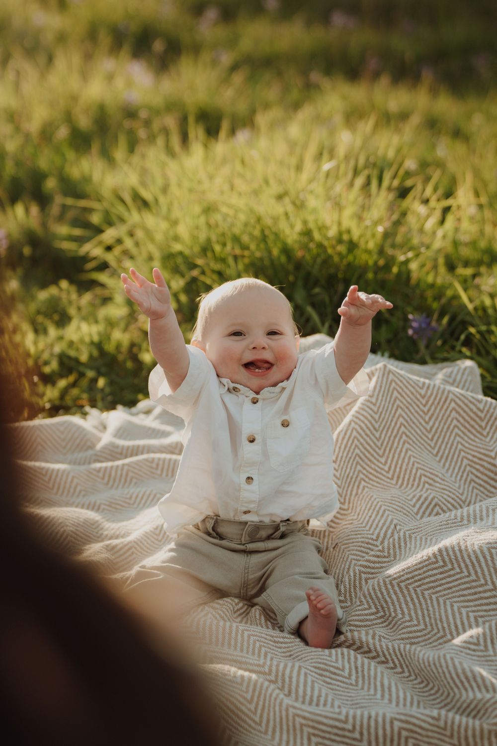 Happy baby sitting on a blanket outdoors, arms raised joyfully, wearing a white shirt and gray pants in golden sunlight.