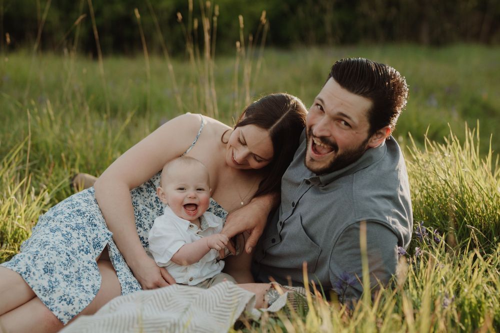 Happy family with baby sitting in green meadow, smiling parents enjoying outdoor portrait session in natural light.