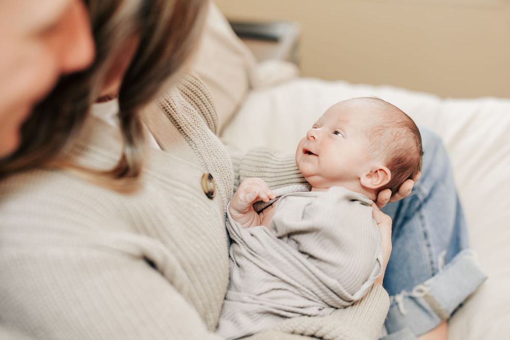 Newborn baby wearing light gray onesie rests in caregiver's arms on white bedding.