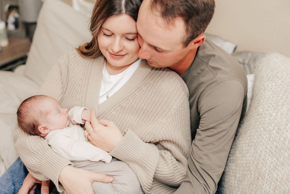 Parents in beige sweater and gray top snuggling with their sleeping newborn baby on couch.