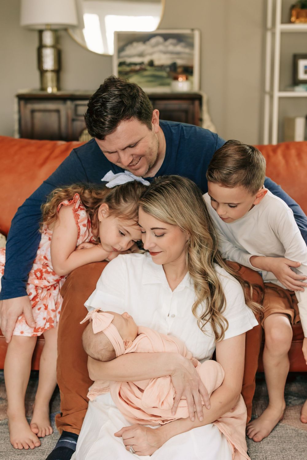 Family gathers on orange couch, sharing tender moment with newborn baby in soft pink blanket at home.