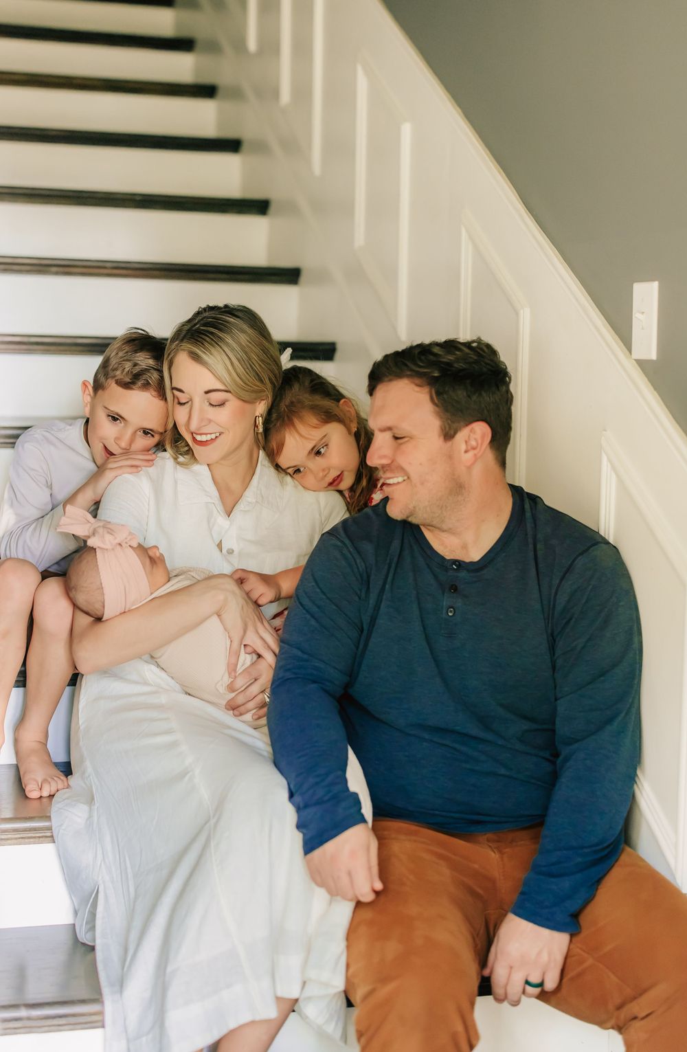 Family sits together on white staircase indoors, wearing casual clothing in warm natural lighting.