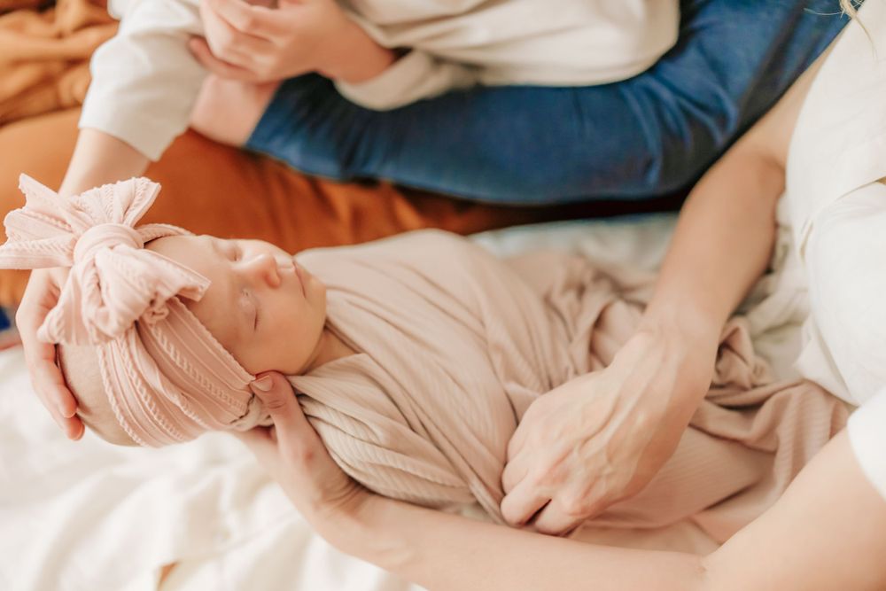Newborn baby wearing soft pink swaddle and headband rests peacefully on white bedding.