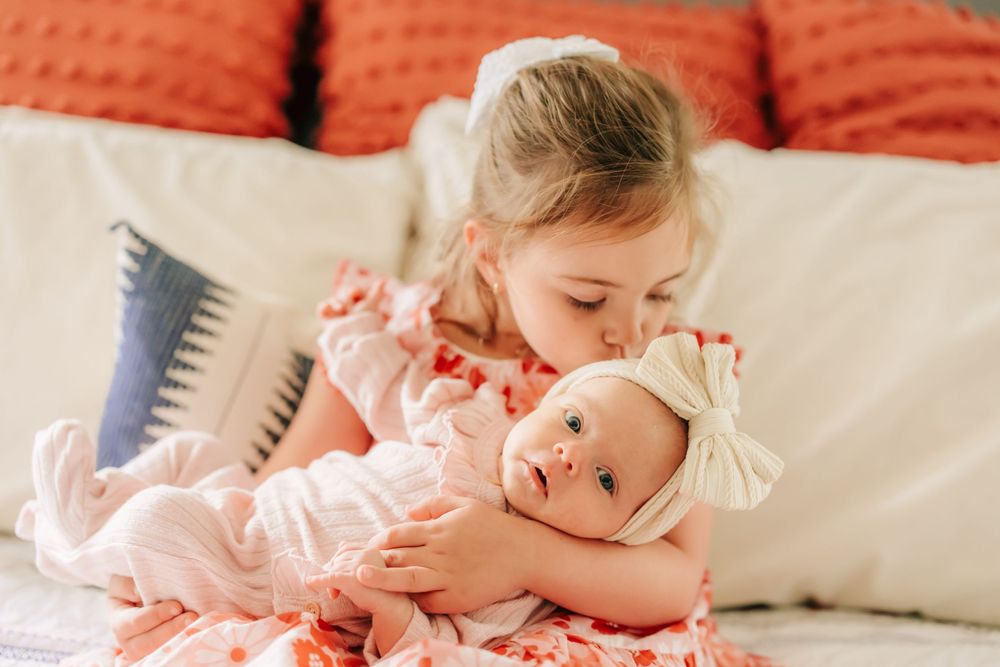 Two siblings in pink dresses share a tender moment on a couch with orange pillows.