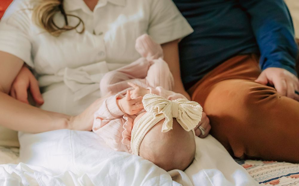 A baby wearing a pink romper with delicate bows lies on a white blanket between two parents.