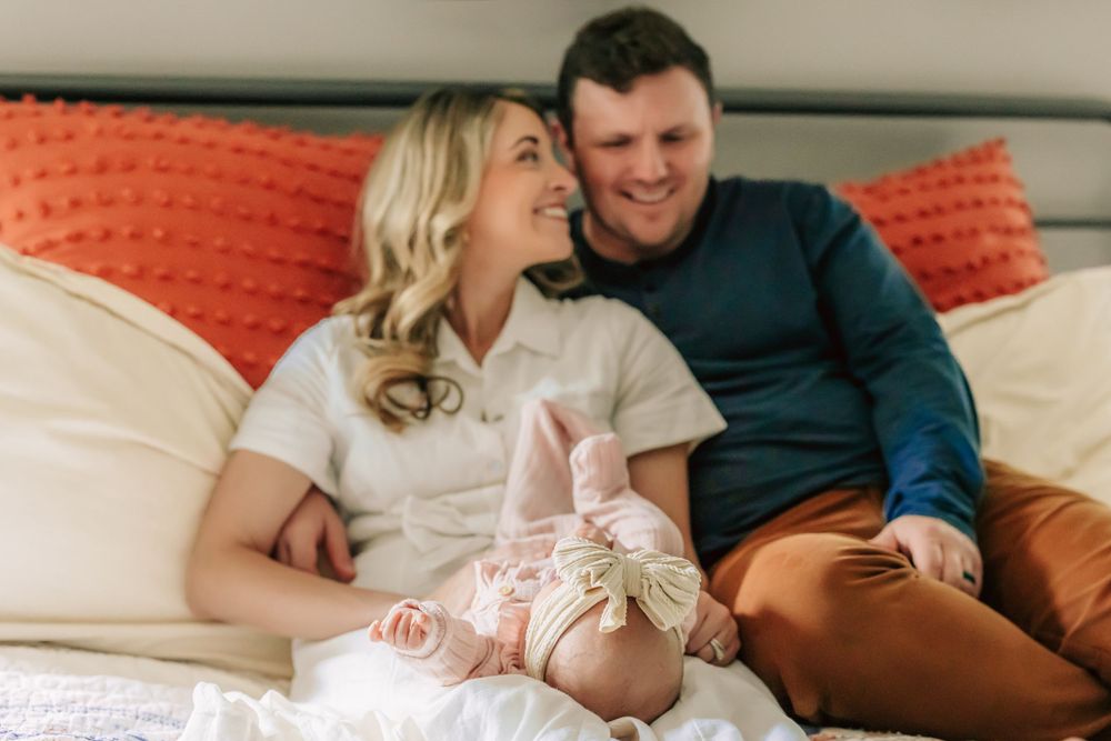 A couple relaxes on a couch with orange throw pillows while smiling and sharing a candid moment together.