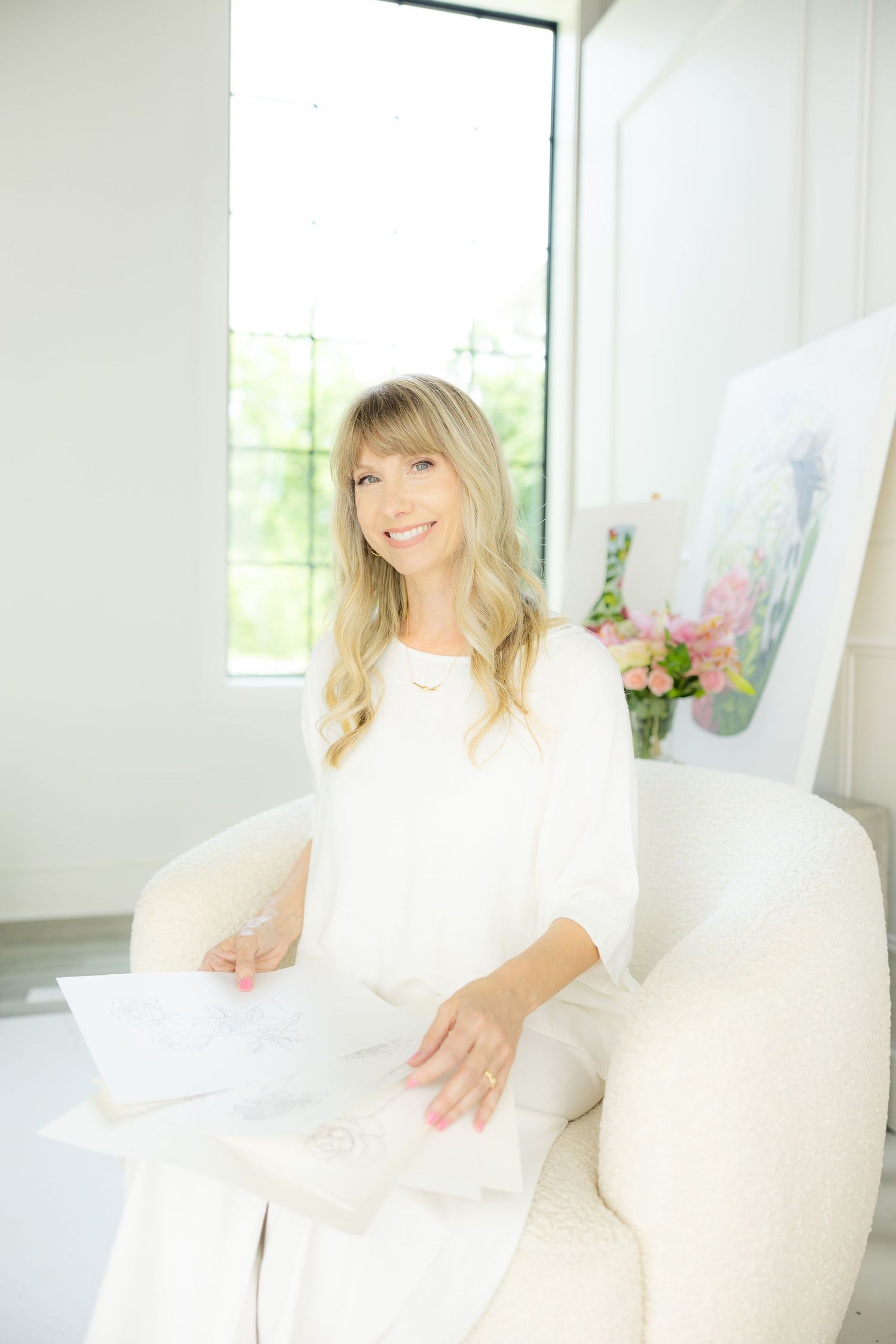 Someone sits in a bright white room on a white couch wearing a white dress with windows and flowers visible in background.