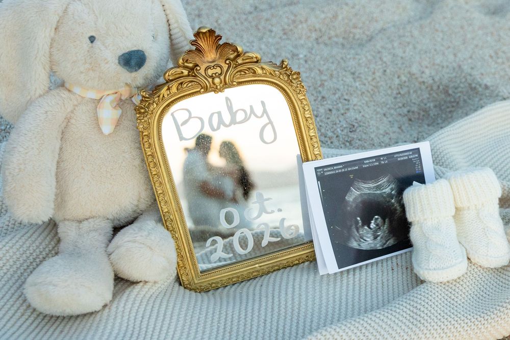 Gold framed mirror with Baby announcement, ultrasound photo, stuffed bunny, and tiny booties on sandy blanket.