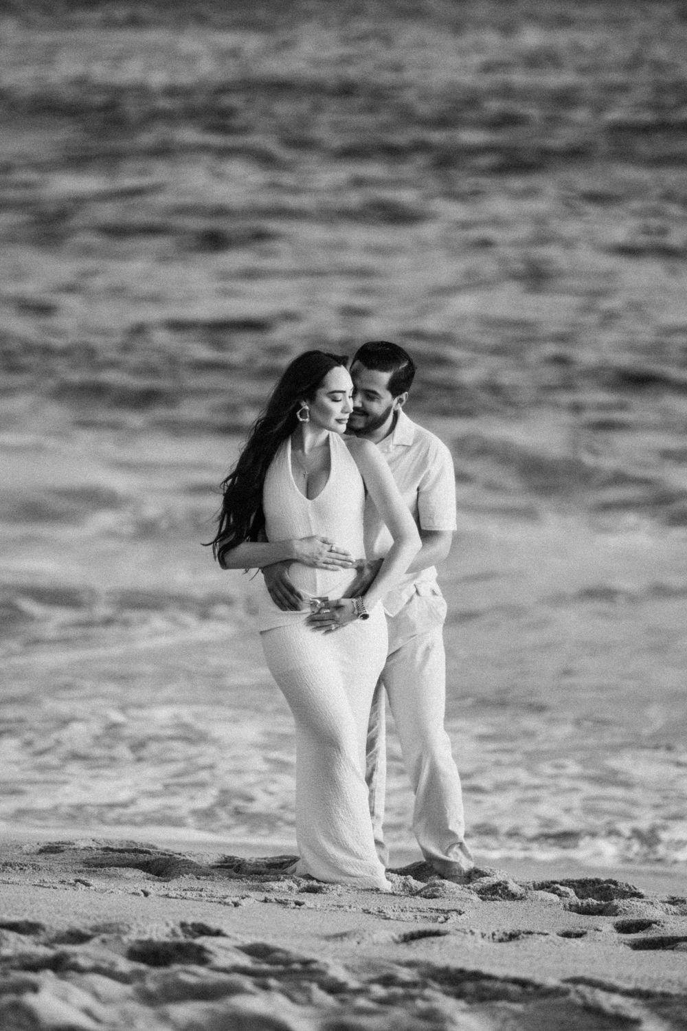 Black and white photo of a couple embracing on a sandy beach with ocean waves in the background.
