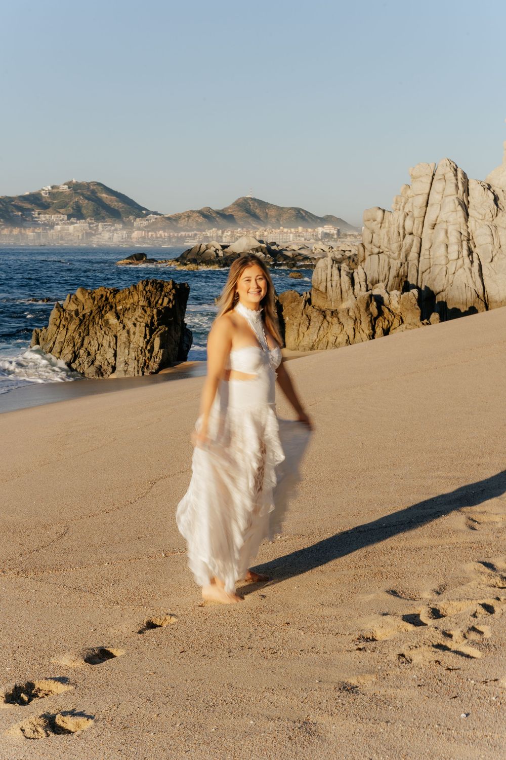A figure in a flowing white dress walks gracefully along a scenic beach with rocky cliffs and ocean views at sunset.