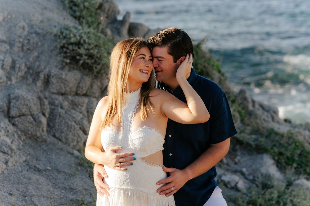 A romantic couple embraces on a rocky cliff overlooking a scenic mountain landscape during sunset.