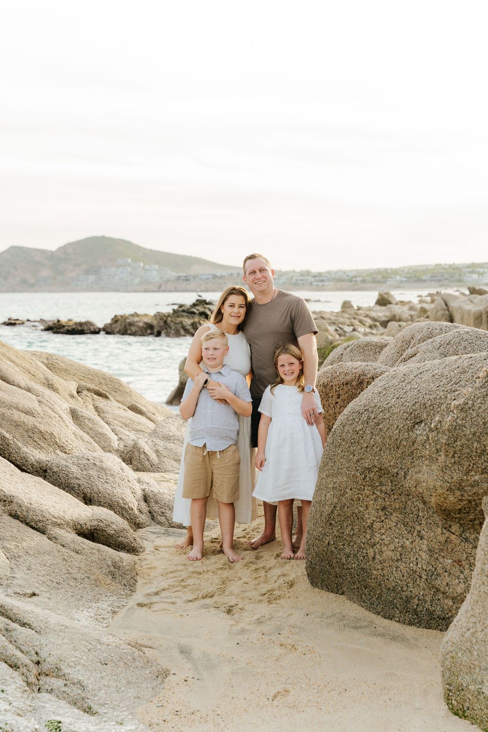 A family stands together on a rocky beach with large boulders and ocean views during sunset.
