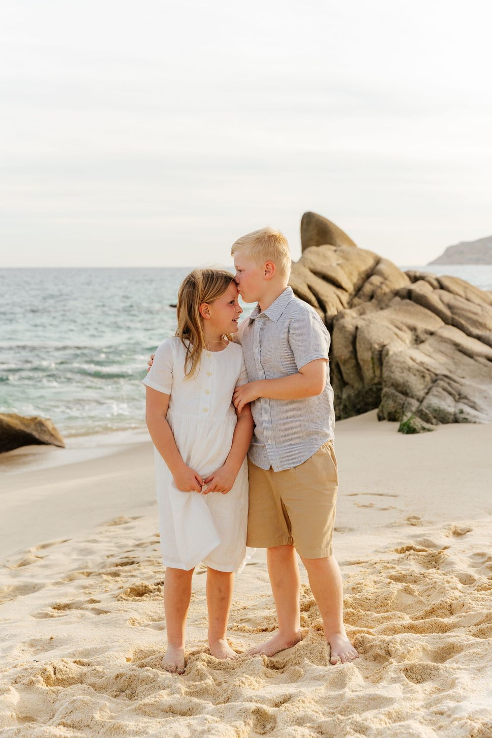 A couple shares a joyful moment together on a sandy beach with large rocks and ocean waves in the background.