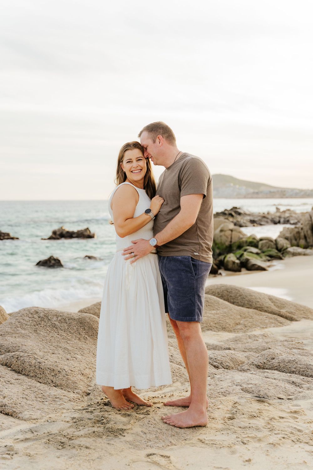 A couple embraces on a scenic beach with large rocks and ocean waves in the background.