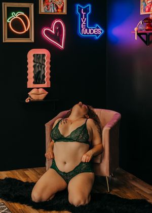 A moody boudoir photo with neon signs on the wall including a heart and vibe symbol, featuring lingerie against dark backdrop.