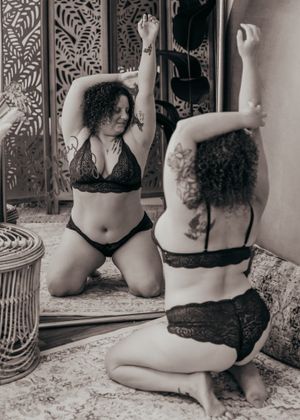 A black and white photo of someone in lace lingerie kneeling and posing with their reflection in an ornate mirror.