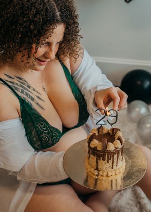 Person in a white cardigan and green lace bralette lighting birthday candles on a chocolate drip cake with festive balloons in background.