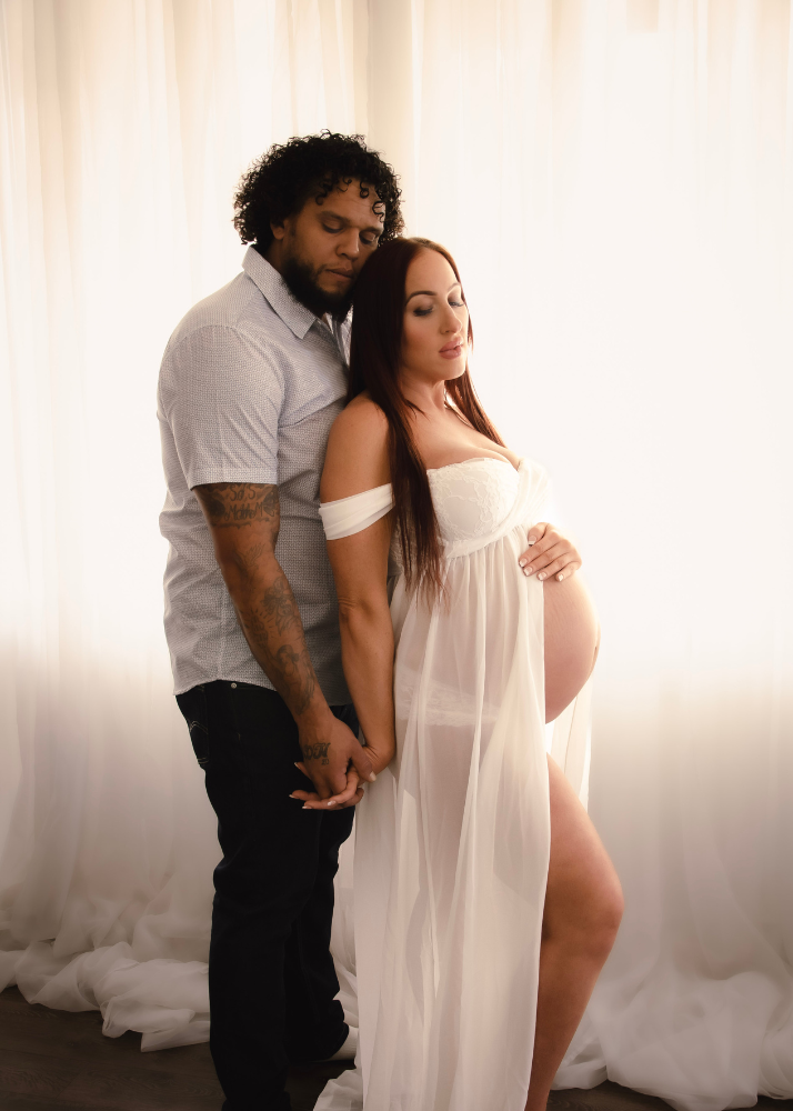 A maternity photo shoot featuring a couple in an intimate pose against a white curtain backdrop.