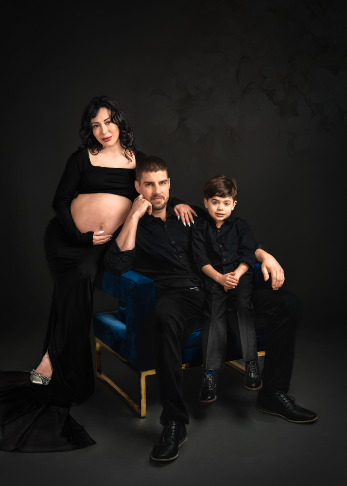 Family portrait with expectant mother in black dress, two others seated on blue chair against dark background.