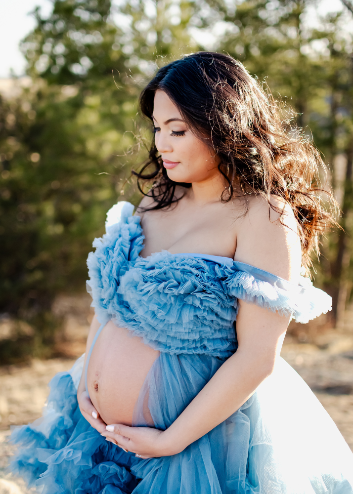 Expecting mother in a flowing light blue off-shoulder maternity gown poses outdoors in golden sunlight.