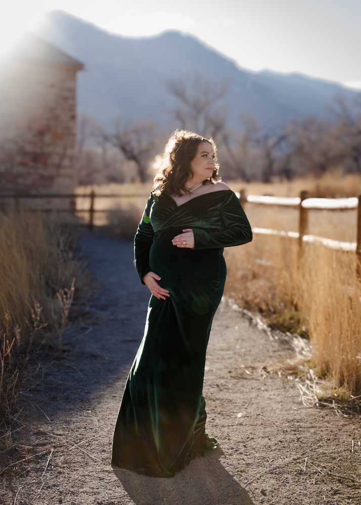 Sunlit maternity portrait of a woman in emerald velvet dress on a rustic mountain path.