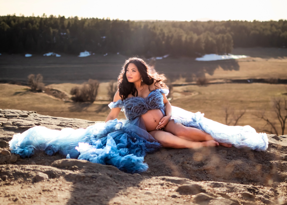 A flowing blue dress creates a dramatic scene against a rocky natural landscape at sunset with rolling hills in the background.