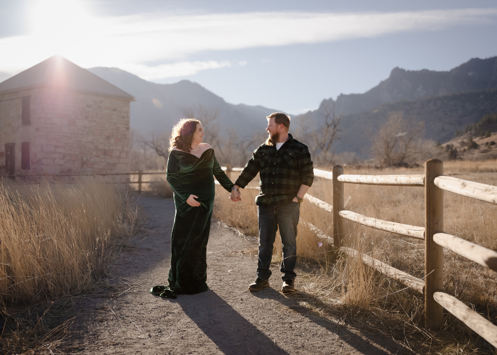 Two people stand on a mountain path with dramatic peaks and sunlight in the background during a winter day.