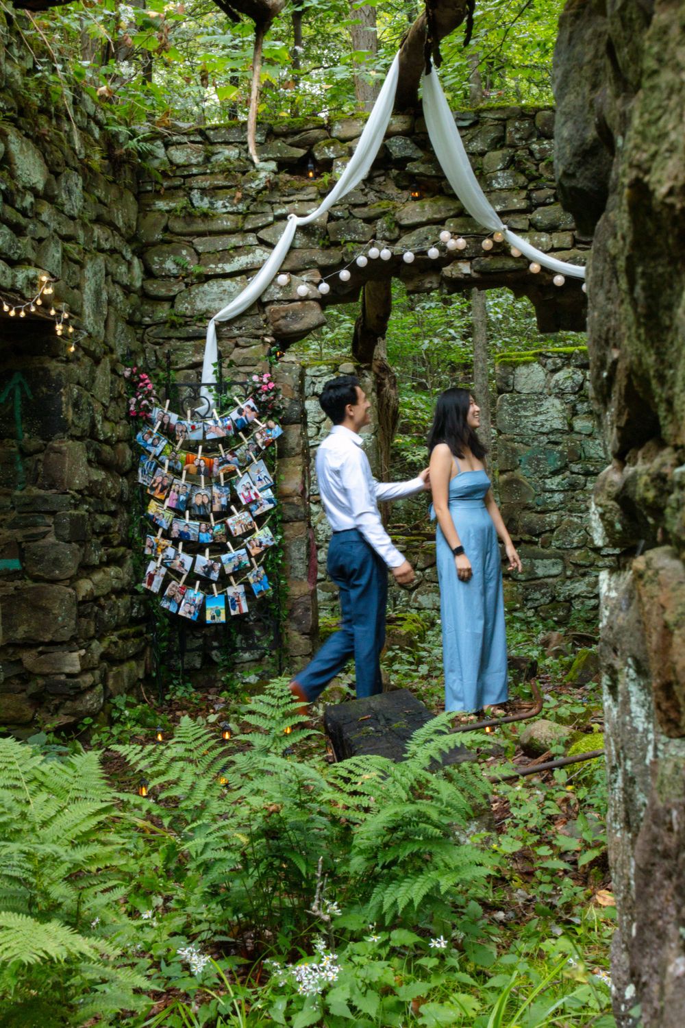 Couple holding hands in decorated stone ruins with photo display and white draping in forest setting.