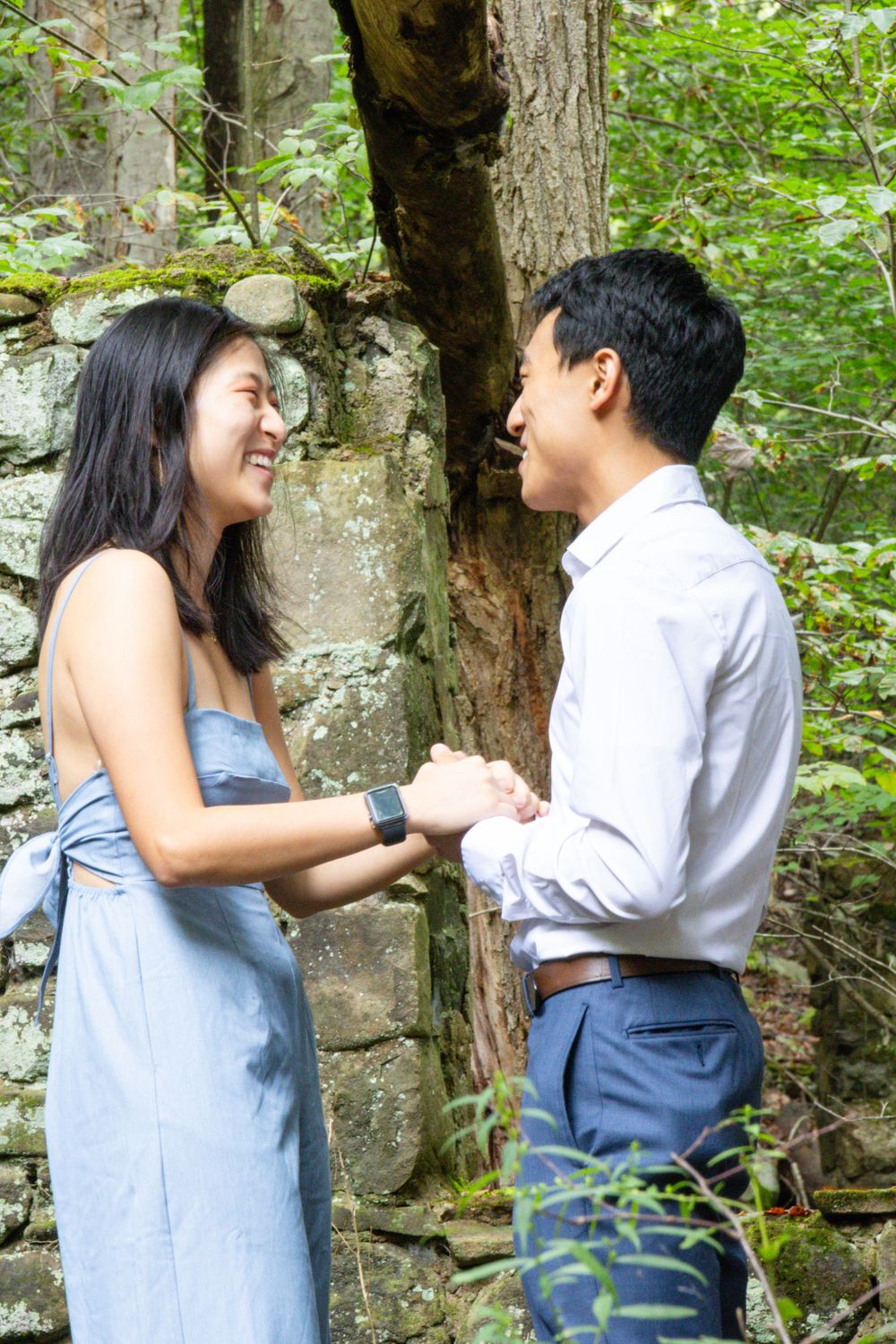 Smiling Asian couple holds hands facing each other beside mossy stone wall in lush green forest setting.