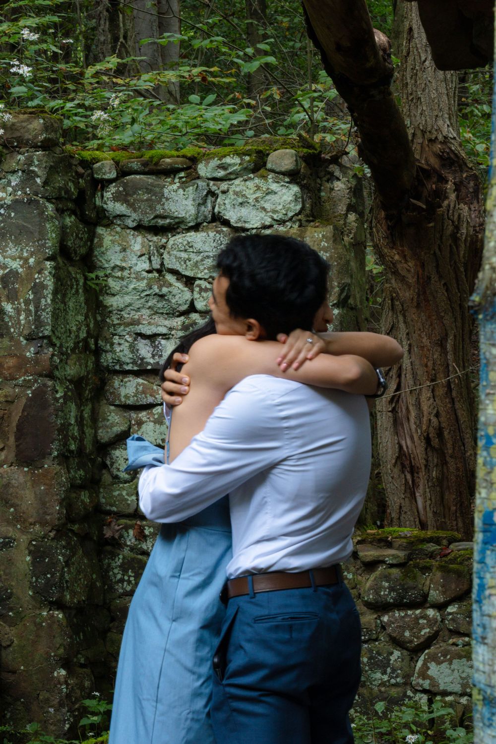 Newly engaged couple sharing a heartfelt embrace against mossy stone wall in woodland setting.