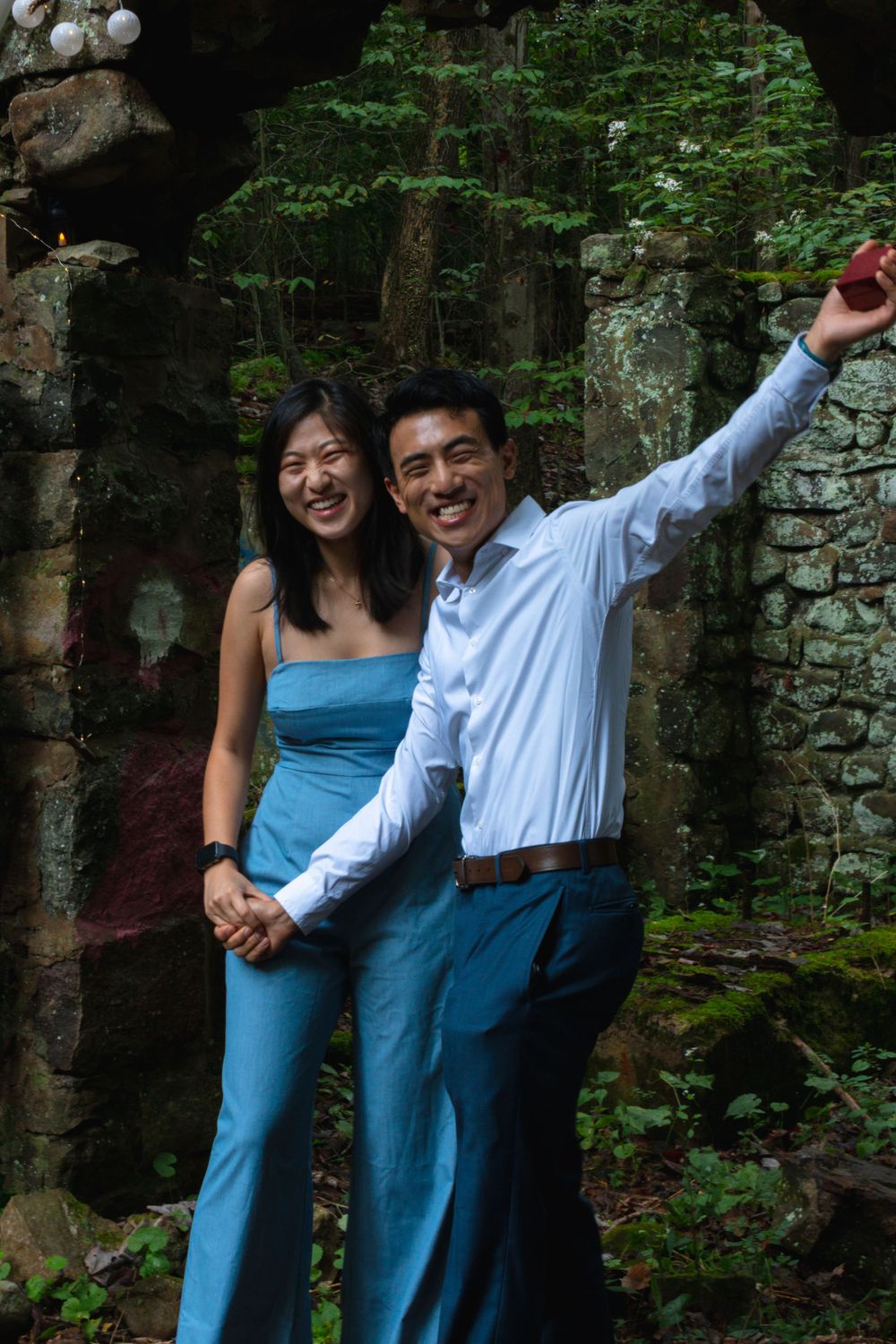 Happy couple celebrating engagement holding ring box in rustic stone ruins surrounded by forest.