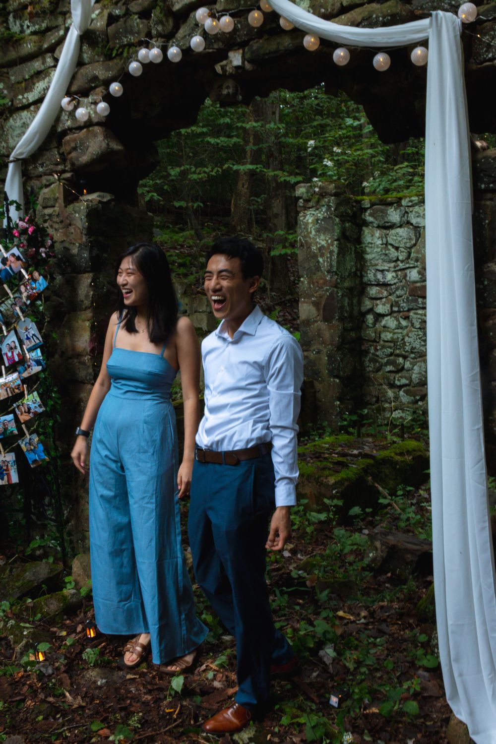 Joyful newly engaged couple laughing together under white draped arch in stone forest ruins.