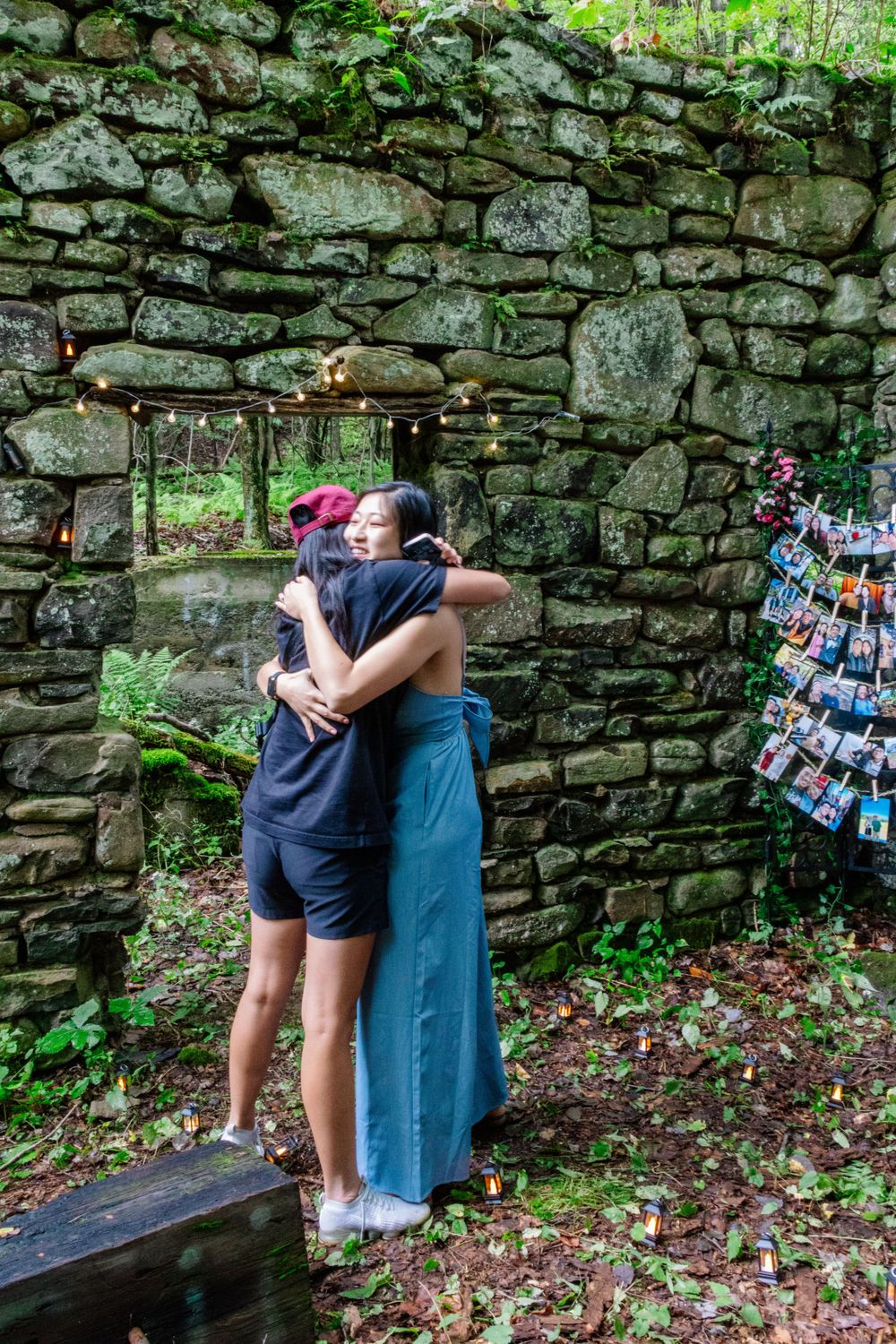 Two women sharing a warm congratulatory hug near stone wall with photo display after proposal.