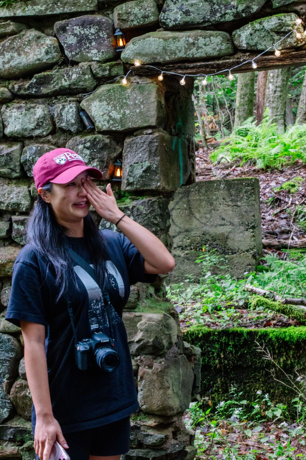 Emotional woman in a pink cap wiping tears near a decorated stone wall with string lights outdoors.