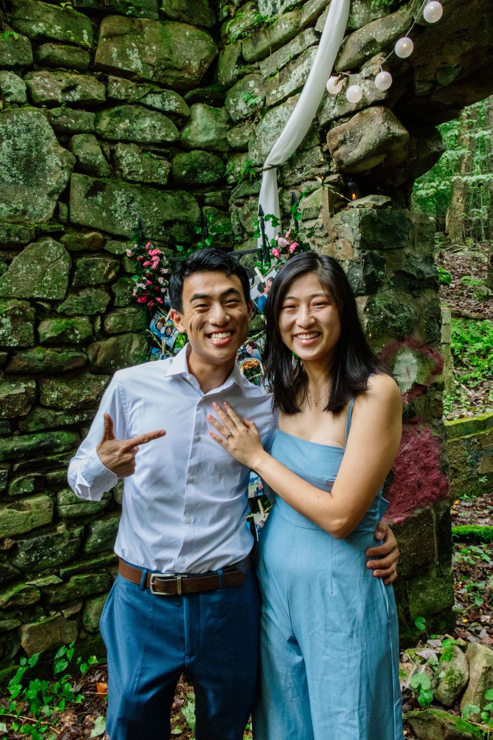 Smiling engaged couple posing together showing off engagement ring against rustic stone wall backdrop.
