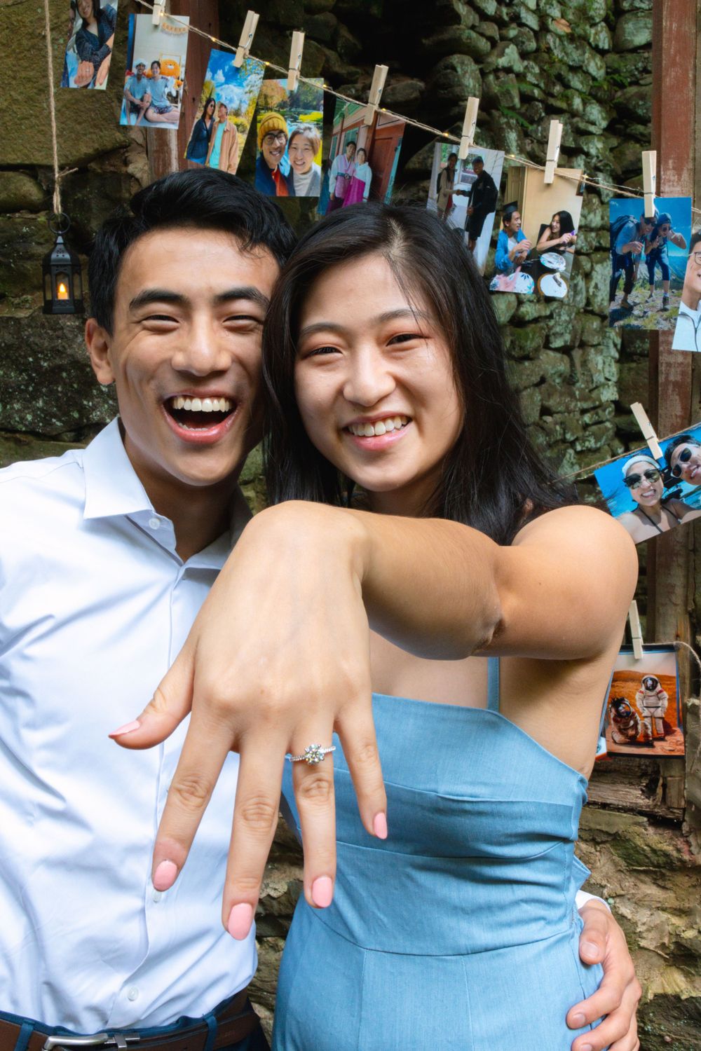 Joyful couple celebrates engagement showing diamond ring with photos hanging on clothesline backdrop.
