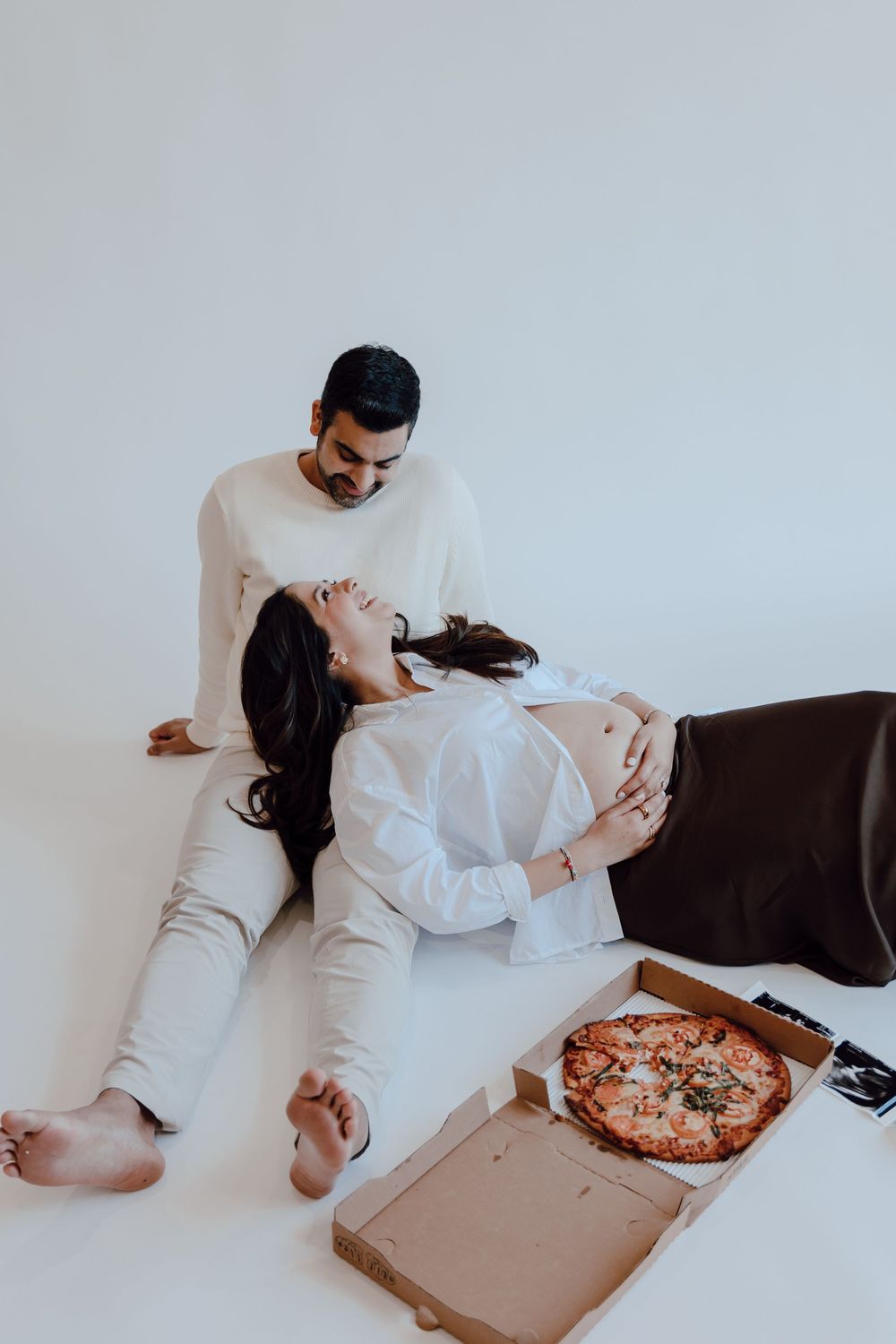 A couple in white loungewear shares a pizza while relaxing on a couch in a minimalist setting.