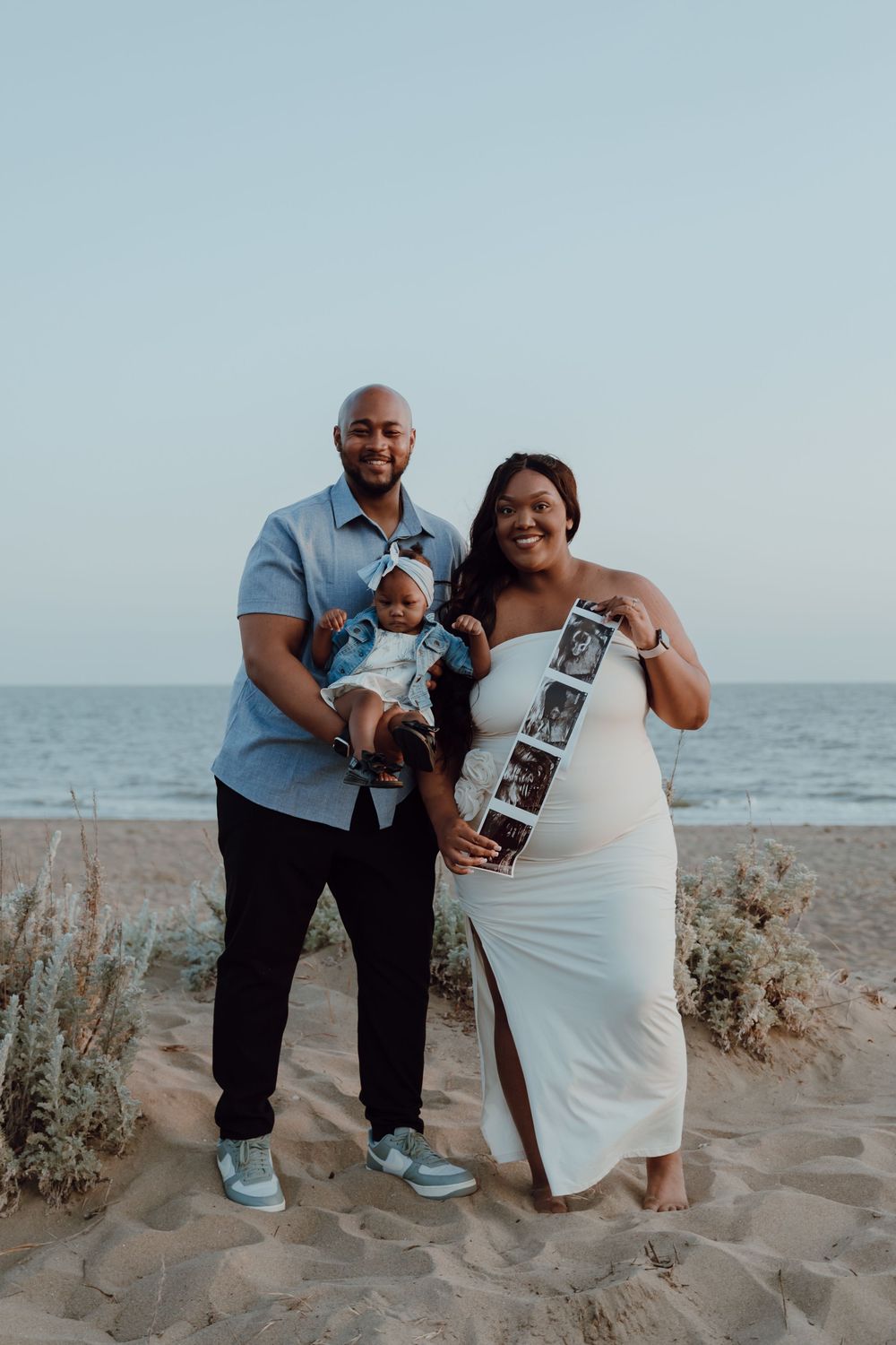 A couple sits together on a sandy beach at sunset holding a sonogram photo.