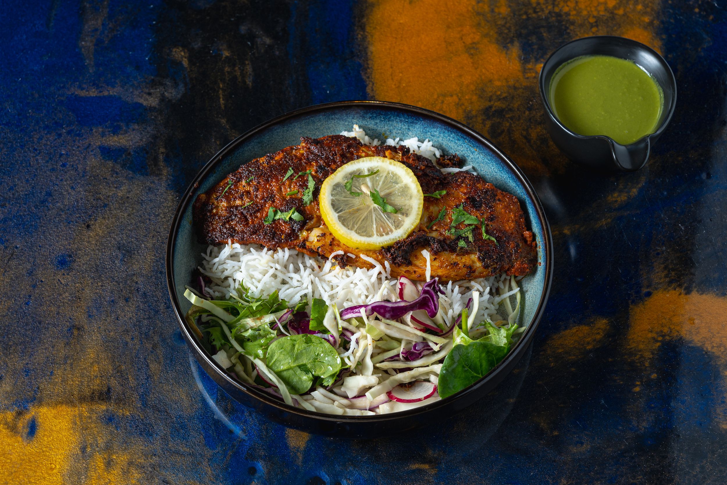 Spiced fish fillet on saffron rice with fresh salad, lemon slice, and green herb sauce in a blue bowl.