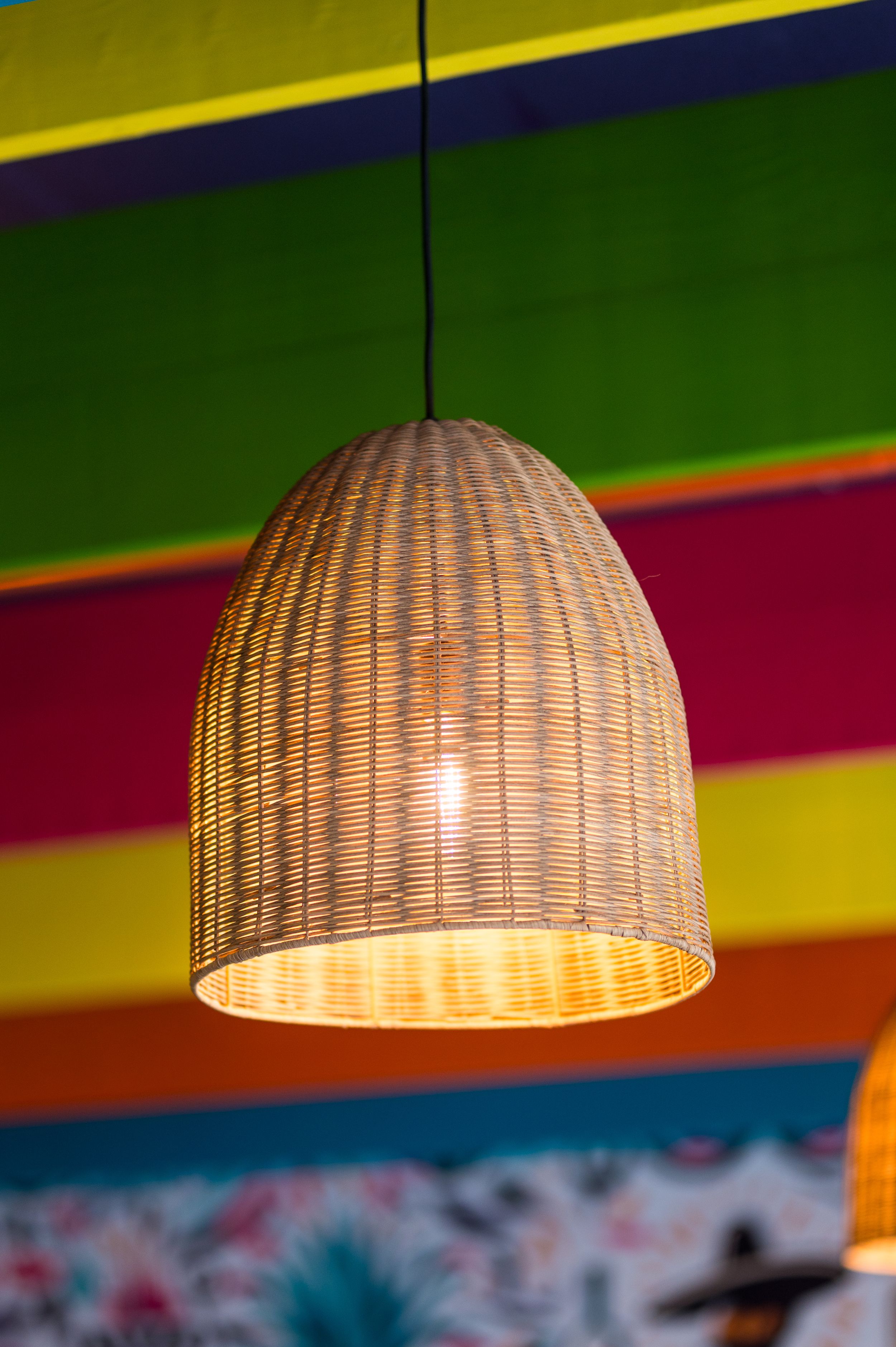 A woven rattan pendant light glowing warmly against a vibrant multicolored striped ceiling in red, green, yellow, and blue.