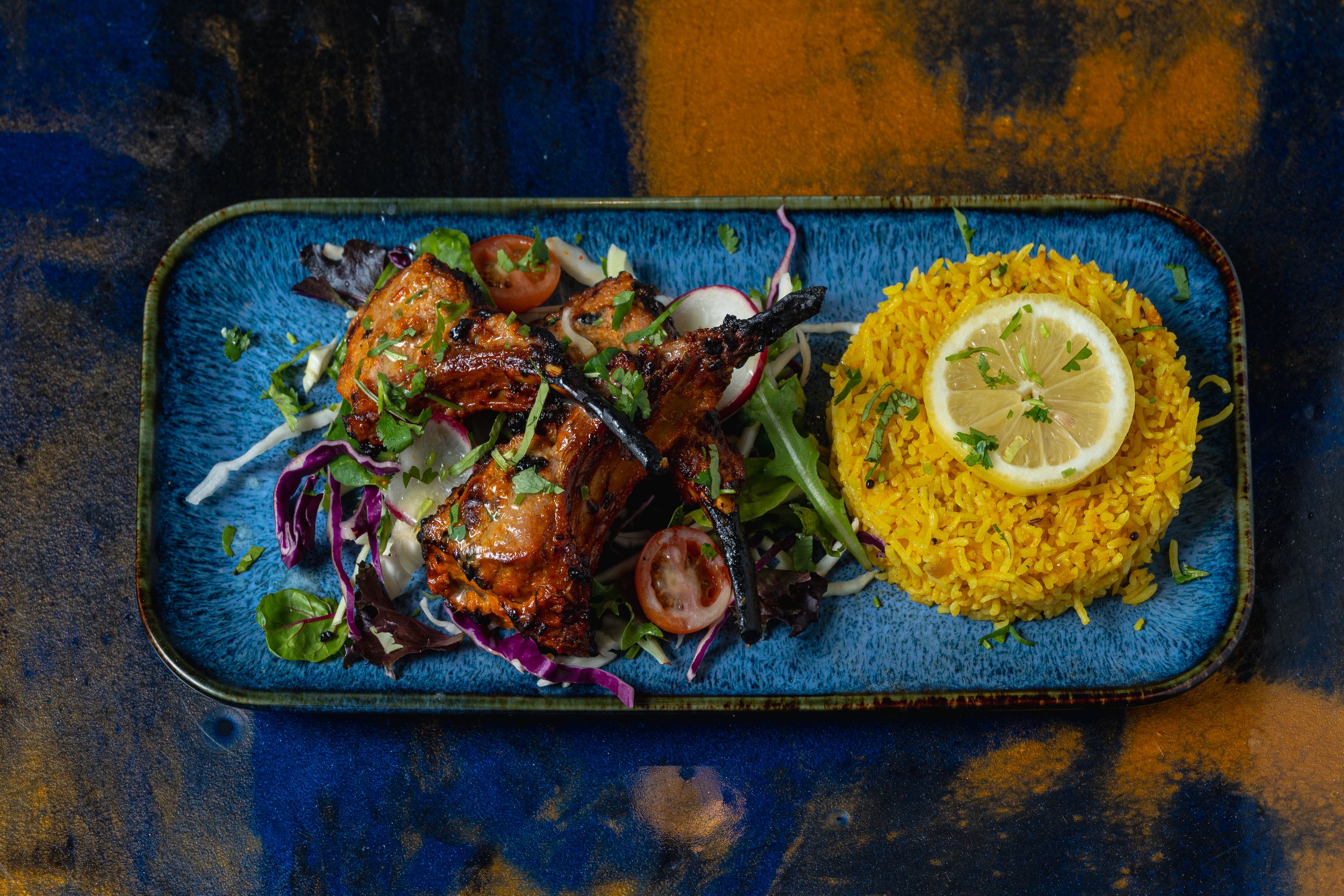 Golden saffron rice and tandoori chicken pieces with mixed salad and lemon slice on a blue ceramic plate.