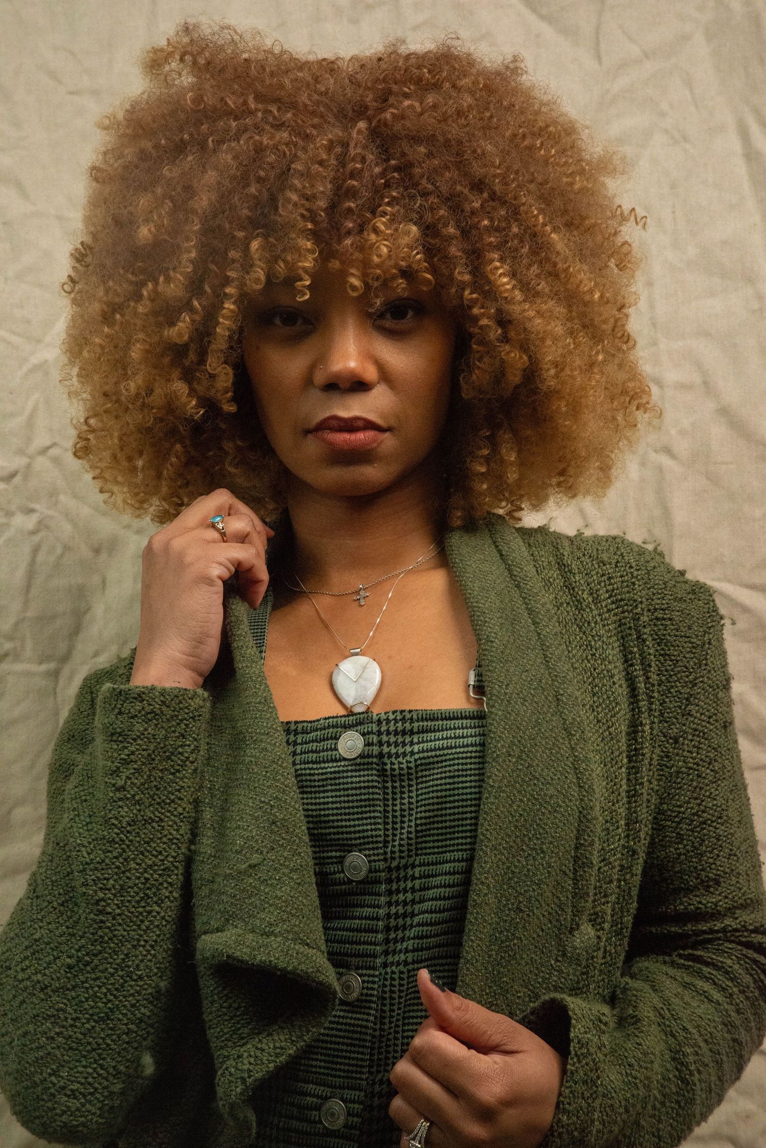 Fashion model wearing olive green cardigan and plaid dress showcasing jewelry and natural curly hair texture.